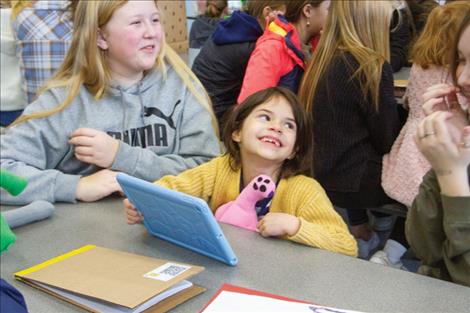 First grader Ruby Epperson smiles up at Zoe Schroeder while holding her new monster stuffie and watching a video based on her drawing. Schroeder and fellow middle schooler Aubrey Wilmar, left, presented Epperson with the book and video they made in their media class for her.