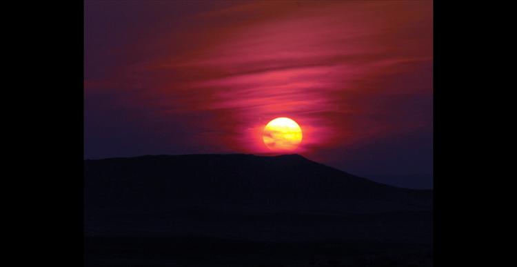 The rising moon glows orange against the background of smoke and early evening light.