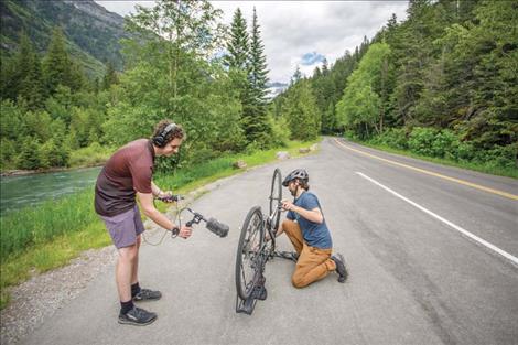 Andrew records, with a shotgun mic, as Michael pedals a bike in a road-side pullout along a creek.