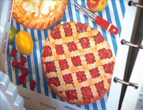 A page in a vintage cookbook shows cherries, a lattice crust cherry pie and and an apple peeler and coreras as well as citurs fruit and a lemon meringue pie. The cookbook is part of the display in the All in Stitches window. 